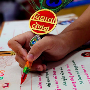 Hand holding a colorful peacock feather pen over a traditional writing board for Chatthi Vidhata Lekhan.