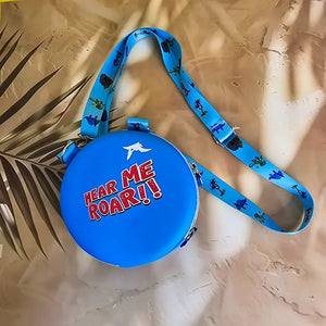 Two colorful, round kids’ sling pouches with attached lanyards are placed on a sandy beige background with a tropical leaf shadow. The left pouch is purple and features an adorable cartoon unicorn with a pink mane, large sparkling eyes, and the name "SHYAMA" written in white. It has a lilac lanyard decorated with candy prints. The right pouch is bright blue with a fierce green cartoon T-Rex dinosaur illustration labeled “DINOSAURS” in bold yellow text. Its lanyard is blue with black dinosaur silhouettes. Bo