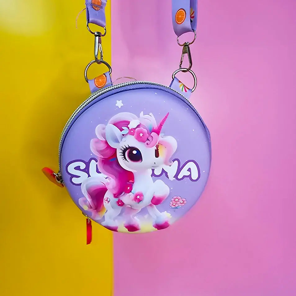 Two colorful, round kids’ sling pouches with attached lanyards are placed on a sandy beige background with a tropical leaf shadow. The left pouch is purple and features an adorable cartoon unicorn with a pink mane, large sparkling eyes, and the name "SHYAMA" written in white. It has a lilac lanyard decorated with candy prints. The right pouch is bright blue with a fierce green cartoon T-Rex dinosaur illustration labeled “DINOSAURS” in bold yellow text. Its lanyard is blue with black dinosaur silhouettes. Bo