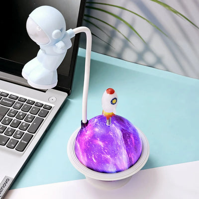 "White astronaut figure desk lamp by Usmile Toys, bending over a glowing purple and blue planet base with a small rocket on top, next to a laptop." www.usmile.toys