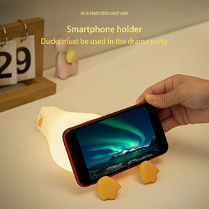 dorable glowing white silicone night lamp for kids, shaped like a little duck lying flat with orange feet and beak, resting on a white surface next to a book. Ideal for a calming bedroom ambiance from uSmile.toys." www,usmile.toys