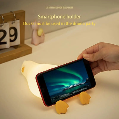 dorable glowing white silicone night lamp for kids, shaped like a little duck lying flat with orange feet and beak, resting on a white surface next to a book. Ideal for a calming bedroom ambiance from uSmile.toys." www,usmile.toys